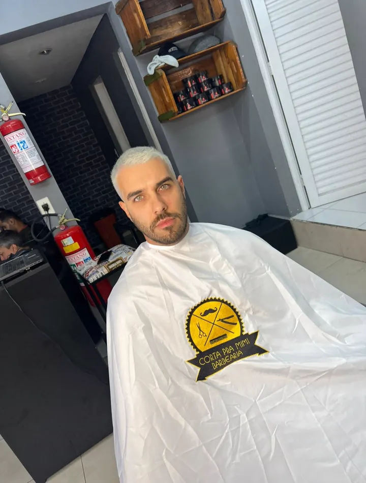 Cliente de cabelo platinado na cadeira, usando a capa branca bordada com o selo da Corta Pra Mim.