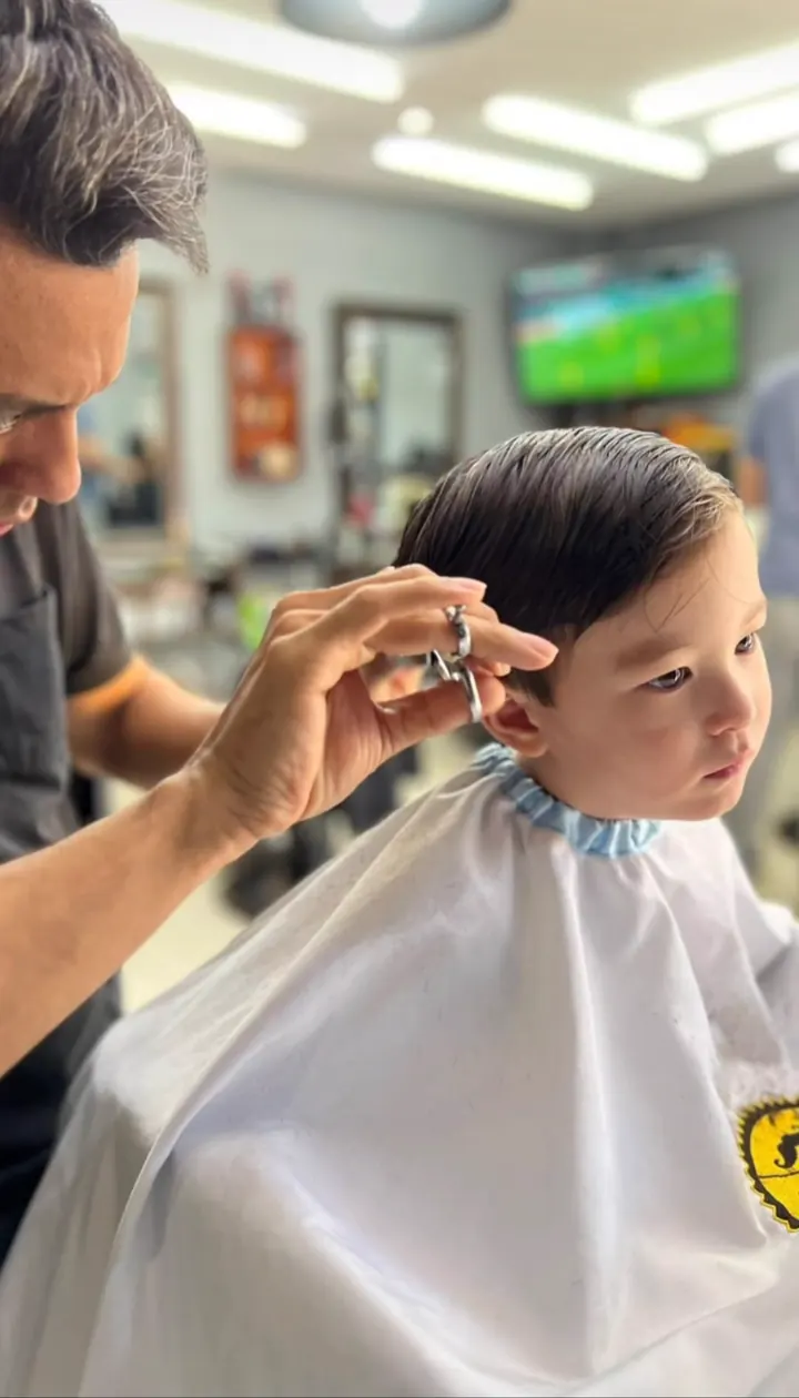 Barbeiro cortando na tesoura o cabelo de um menino pequeno, de capa branca.